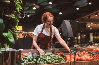 Young smiling seller in apron standing behind counter with vegetables happily working in modern supermarket