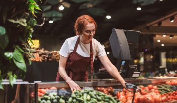 Young smiling seller in apron standing behind counter with vegetables happily working in modern supermarket