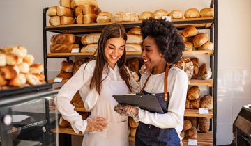 Two beautiful young bakers at the bakery. Shot of a young woman showing her colleague something on her clipboard while they stand in their bakery shop. Smiling young bakers at the bakery shop